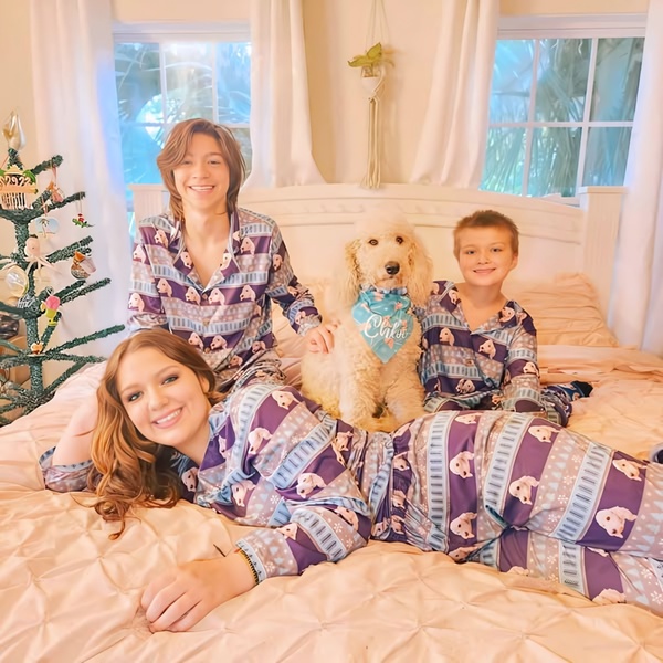 A high-quality lifestyle image of a family and their dog lounging on a bed wearing matching, personalized holiday-themed pajamas.
