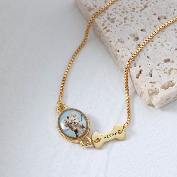 A close-up product shot of a gold necklace featuring a custom pet photo pendant and a small bone-shaped name tag.