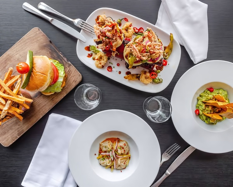 A high-angle, top-down photography shot displaying an assortment of gourmet dishes including a burger, dumplings, and fresh vegetable sides.