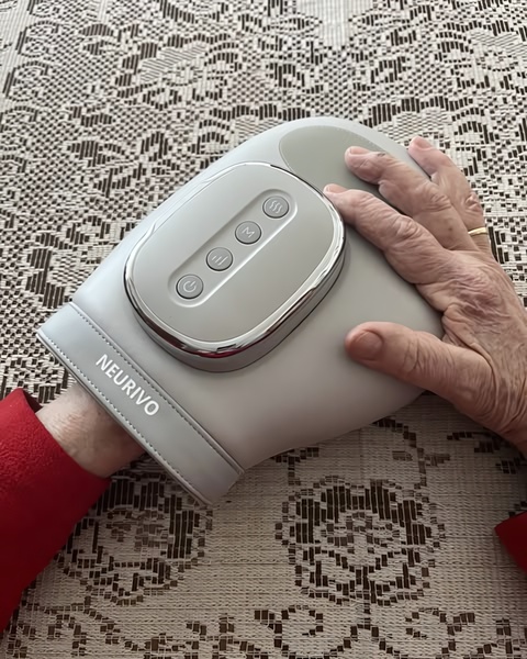 A close-up, top-down shot showing an elderly person's hand resting on a knee massager device placed on a neutral table setting.