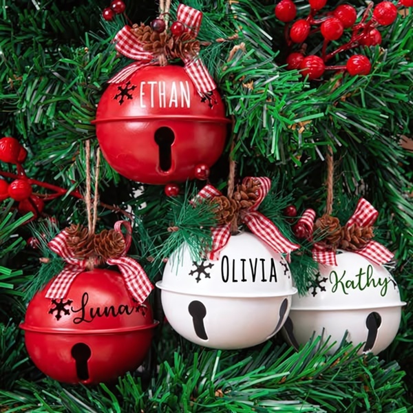 A close-up, festive image of personalized red and white holiday jingle bell ornaments hanging on a Christmas tree with pine cones and berries.