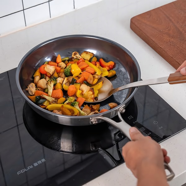 A close-up, high-angle shot of a person using a spatula to stir-fry a mix of vegetables in a high-quality stainless steel pan on an induction cooktop.