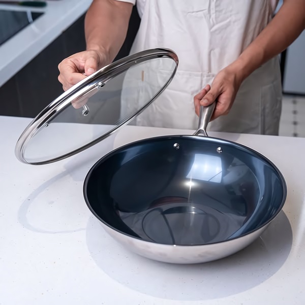 A high-quality product shot of a stainless steel frying pan with a glass lid, set on a bright, minimalist kitchen countertop.