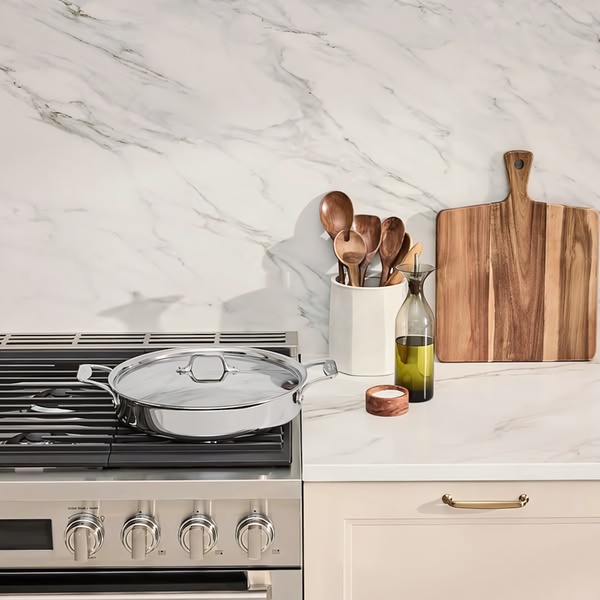 A high-quality, bright, and airy photograph featuring a stainless steel skillet on a stovetop, surrounded by marble textures and wooden kitchen tools.