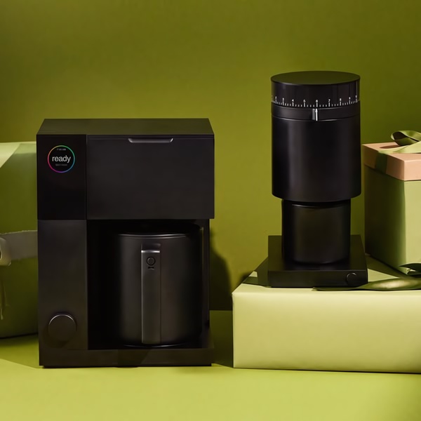 A sleek, black coffee grinder and matching coffee maker arranged elegantly next to gift-wrapped presents on a solid muted green background.