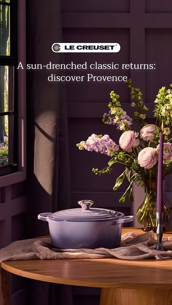 A high-end product hero shot featuring a purple enameled cast iron pot placed on a wooden table, styled with fresh flowers in a home interior setting.
