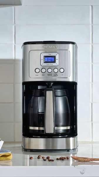 A high-fidelity studio shot of a stainless steel coffee maker on a clean white kitchen counter, surrounded by coffee beans to establish the mood.