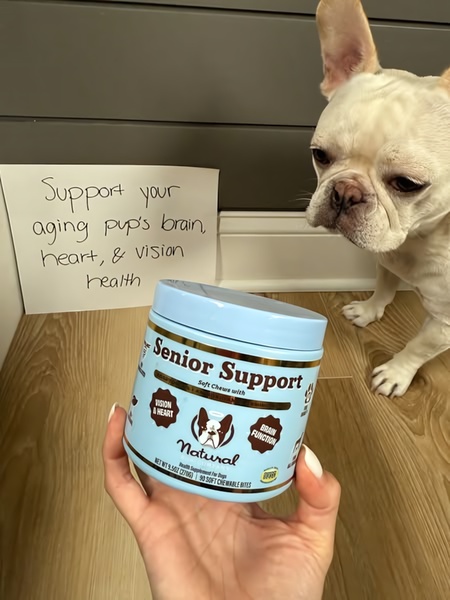 A close-up, hand-held shot of a dog supplement container positioned next to a dog, paired with a handwritten benefit sign.