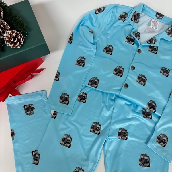 A flat-lay view of light blue pajamas featuring a repeating photo pattern of a pet, arranged neatly with festive holiday decor nearby.