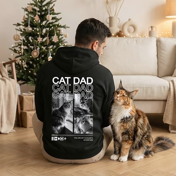 A man wearing a black Cat Dad hoodie sits on the floor next to his pet cat near a decorated Christmas tree.