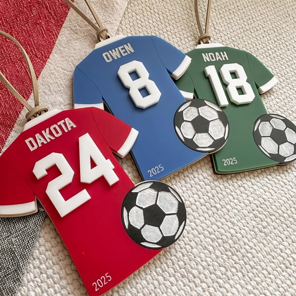 A high-angle studio shot of three custom-colored, jersey-shaped soccer ornaments featuring names, player numbers, and the year 2025.