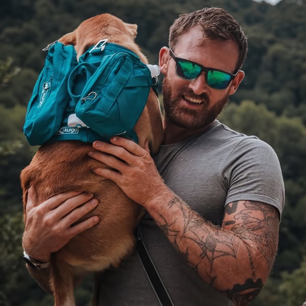 A close-up shot of a man wearing a teal tactical-style backpack on his dog while hiking in a natural, outdoorsy environment.