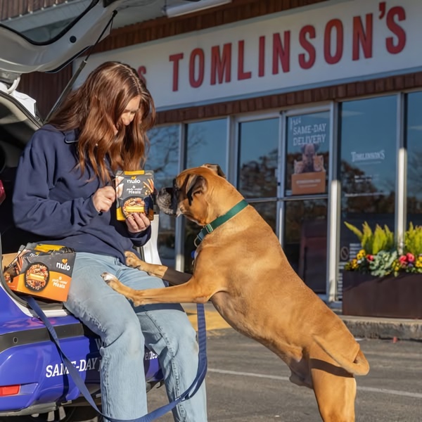 A pet owner interacts joyfully with their boxer at a storefront, clearly displaying the dog food product in a relatable, real-world setting.