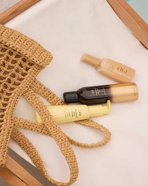 A top-down, high-aesthetic product shot featuring three Oribe hair serum bottles arranged naturally in a straw tote bag on a neutral surface.