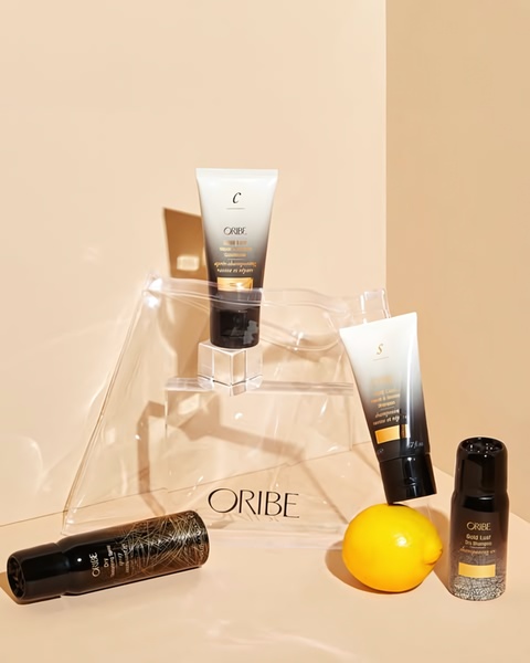 A high-end, studio-lit display of Oribe hair care bottles and a lemon, organized in an asymmetrical, minimalist composition on a soft beige background.