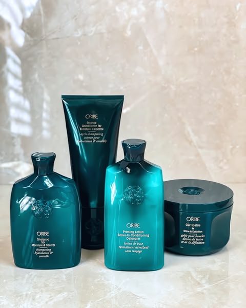 A clean, high-end studio shot of four Oribe hair care bottles arranged in a balanced composition against a neutral marble backdrop.