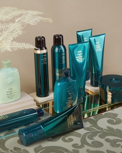 A high-end product display showcasing a collection of Oribe hair care bottles and tubes arranged on a reflective surface against a neutral background.