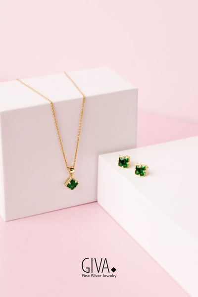 A high-end, clean studio shot of a gold necklace and matching square-cut green stone earrings resting on minimalist white pedestals.