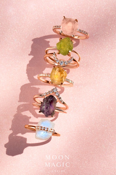 A vertical product hero shot featuring a vertical stack of five unique crystal rings on a soft, glittery pink background.