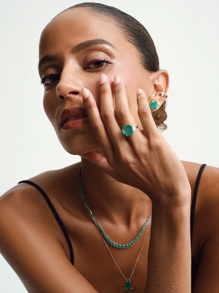 A close-up portrait of a model showcasing an emerald ring and matching necklace in a clean, minimalist studio setting.