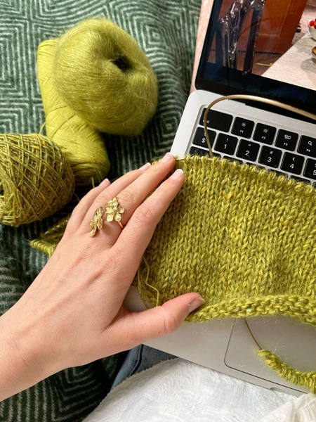 A close-up, first-person perspective of a user knitting with vibrant lime green yarn while working on a laptop, emphasizing a cozy and productive hobbyist aesthetic.