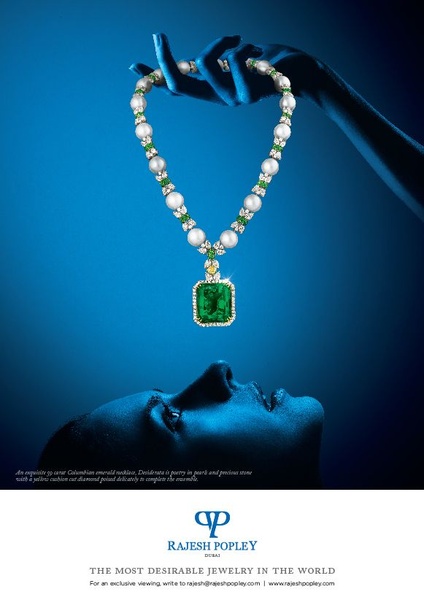A striking high-end jewelry creative featuring a 99-carat Columbian emerald necklace suspended beneath a human silhouette, set against a deep blue background.
