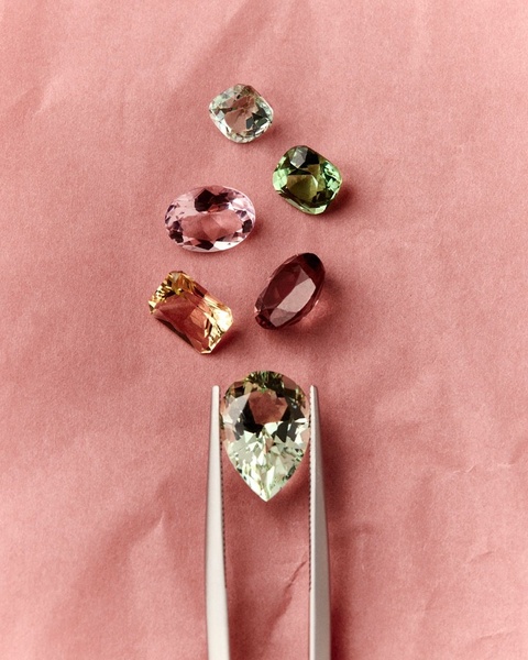 A close-up view of a high-quality gemstone being held by tweezers, surrounded by a curated selection of loose, colorful stones on a textured pink background.