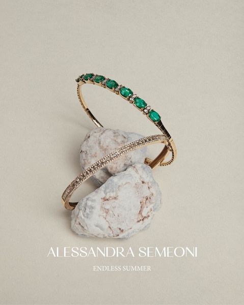 A high-end editorial product shot featuring two gold-toned bracelets resting on natural stones, emphasizing an airy, premium summer aesthetic.