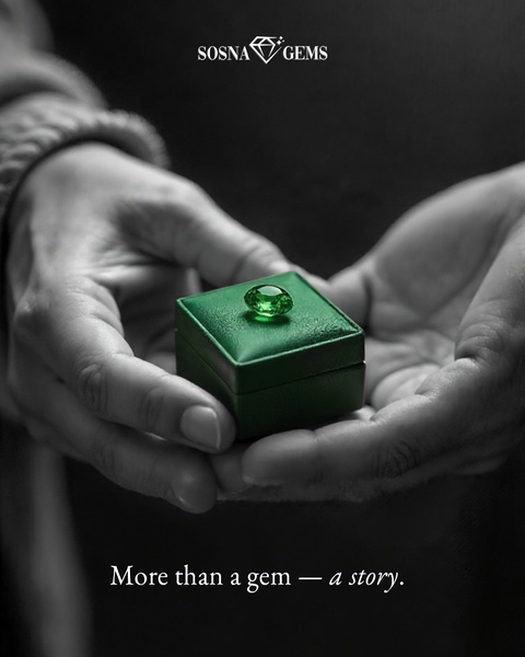 A close-up, black and white image features hands gently holding a small box containing a single, vibrant green gemstone.