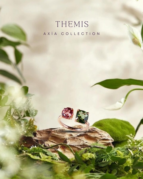 A close-up product shot of a gold ring with pink and green stones, positioned on a natural mossy stone surrounded by white flowers.