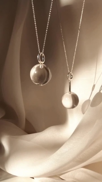 A high-end product hero shot featuring two pendant necklaces suspended against draped, neutral-toned fabric, emphasized by warm, natural sunlight.