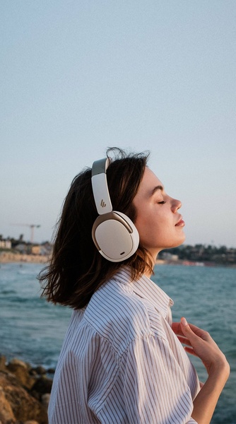 A close-up, side-profile shot of a woman wearing modern white wireless headphones in a calm, outdoor coastal environment at dusk.