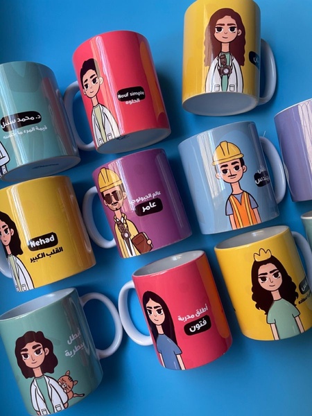 A high-angle, top-down shot showcasing a collection of personalized mugs featuring unique illustrated characters and Arabic name labels on a solid blue background.