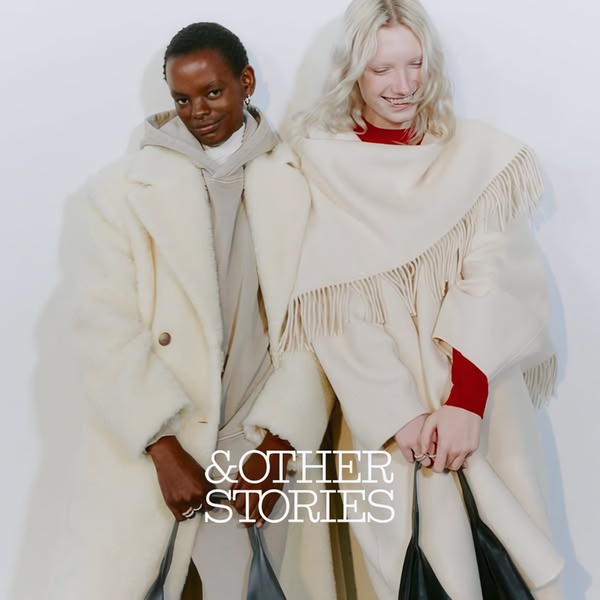 Two models showcasing a cozy off-white wool coat and fringe scarf in a high-fashion, minimalist studio setting.