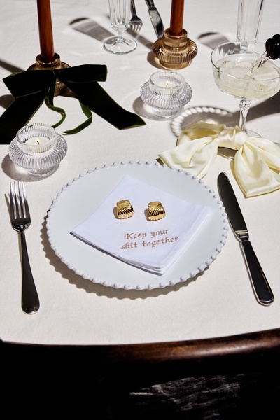 A high-end product shot featuring a pair of gold earrings placed on a white napkin with the phrase Keep your shit together, set within a formal, candlelit dinner table environment.