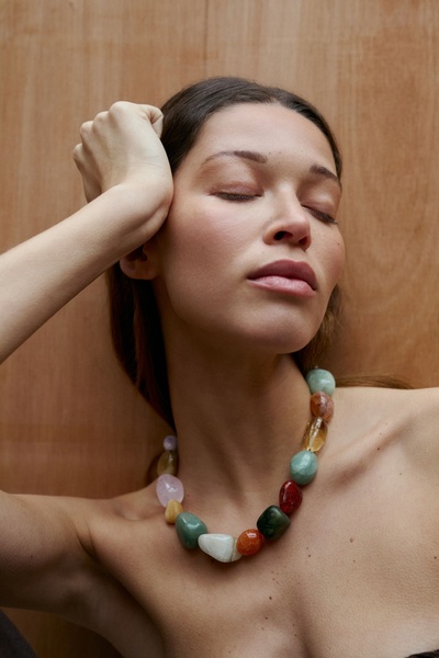 A close-up portrait of a model wearing a statement necklace with multi-colored smooth stones against a simple wooden background.