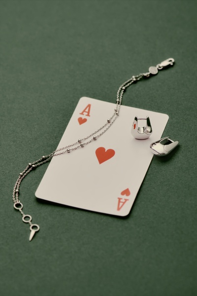 A high-end product shot featuring silver hoop earrings and a delicate chain draped over an Ace of Hearts playing card on a dark green surface.