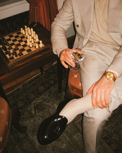 A high-angle, lifestyle-focused image featuring a pair of premium leather loafers, a man in a tailored suit, and a glass of scotch next to a chess board.