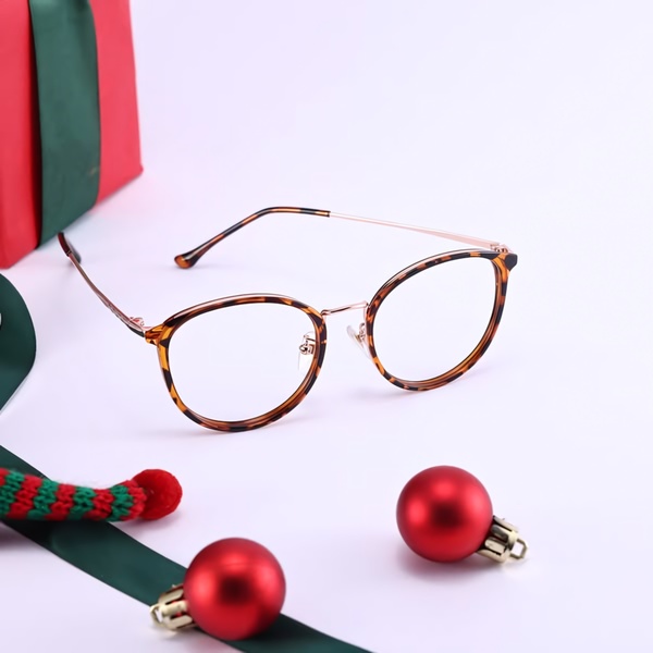 A clean, high-contrast studio shot of tortoise shell glasses surrounded by holiday ornaments, emphasizing a festive and premium brand aesthetic.