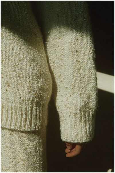 A close-up, high-detail image of a cream-colored textured knit sweater, emphasizing material quality through natural side-lighting.