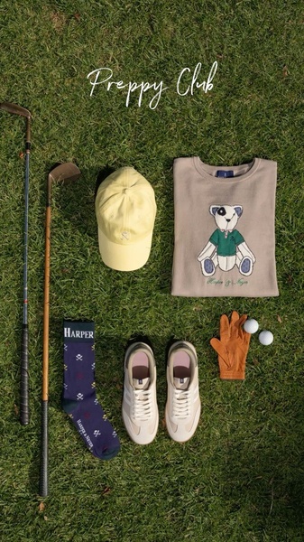A high-angle flat lay composition displaying a curated golf-inspired outfit, including a bear graphic sweatshirt, cap, socks, and golf clubs on a grass surface.