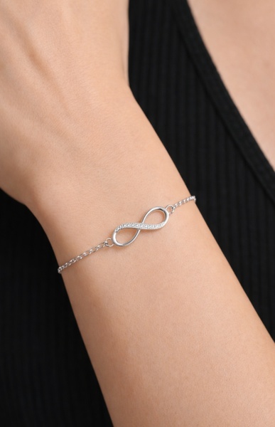 A close-up, high-quality image of a delicate infinity-symbol bracelet worn on a person's wrist against a solid dark background.