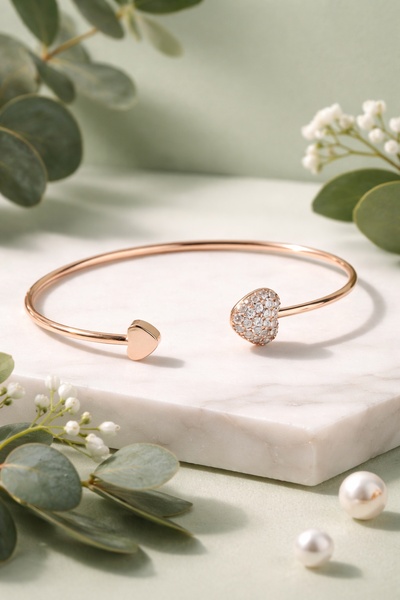 A high-end studio hero shot of a rose gold heart-shaped cuff bracelet set against a soft marble and botanical backdrop.