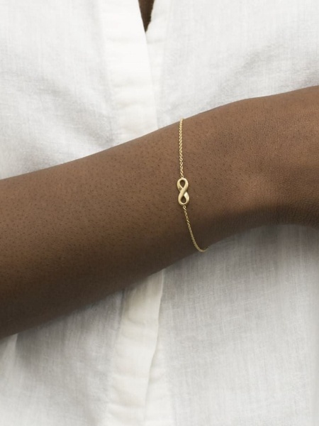 A close-up shot of a delicate gold infinity bracelet on a person's wrist against a soft white linen background.
