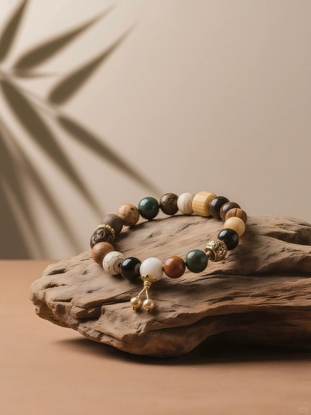 A multi-material beaded bracelet is elegantly displayed on a piece of raw driftwood, set against a neutral background with a soft palm leaf shadow overlay.