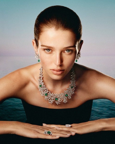 A luxury-focused portrait featuring a model wearing an ornate diamond and emerald necklace and rings against a dark water backdrop.