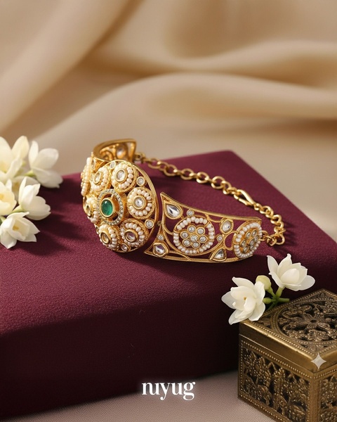 A close-up studio shot of a gold, pearl, and emerald bracelet displayed on a deep burgundy surface with white jasmine flowers.