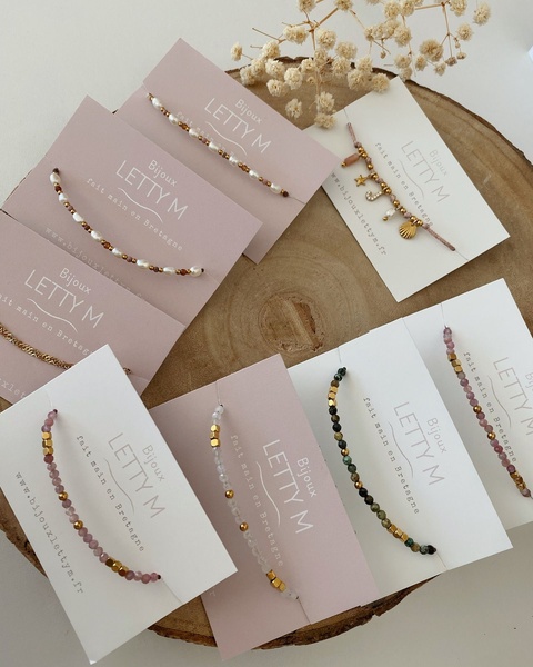 A flat lay image displaying multiple handmade bracelets on individual branded cards against a natural wood background, accented with dried foliage.