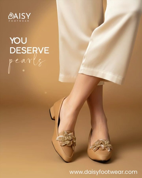 A close-up, high-quality photograph of a pair of beige embellished flats worn with white trousers, focusing on the product's intricate details.