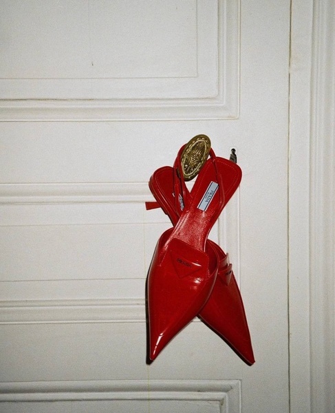 A high-fashion, minimalist product shot featuring red slingback heels hung from a classic brass door handle against a neutral white door.